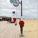 Cloudy Day on Beach Boardwalk with Clock and Flag Beach boardwalk with clock, flag, dunes, and people enjoying a cloudy coastal day.