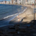 Tranquil Beachfront With City Skyline and Ocean Waves Sunny beach with city skyline, ocean waves, and relaxing tourist enjoying the seaside.