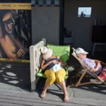 Relaxing Beachside Couple With Vintage Poster Backdrop Elderly couple relaxing on beach deck chairs with vintage Suzy Solidor poster backdrop.