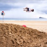 Whimsical Leap Over Sand Mound on Beach Person jumping over sand mound on beach with red ball, ocean waves, and island background.
