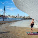 Meditation by the Ocean With City and Sculpture Backdrop Man meditating on beach deck with ocean, city skyline, and coastal sculpture backdrop.