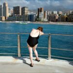 Woman in Swimsuit Admiring Ocean View from Pier Woman in black swimsuit gazing at ocean from pier with beach and city skyline.