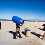 Men Enjoy Quirky Day at Sunny Beach Men on sandy beach with blue barrel, ocean waves, and sailboats in background.