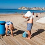 Sunny Beach Day With People Enjoying the Shore Sunny beach scene with people relaxing, ocean waves, umbrellas, and coastal town background.