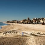 Solitary Sand Art Spirals on Peaceful Beach Shore Person creating spiral sand art on peaceful beach with ocean waves and clear blue sky.
