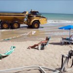 Beach Sunbathers Overlook Truck Maintenance by Shoreline Beach maintenance truck driving near sunbathers on sandy shore with ocean waves.