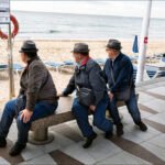 Elderly Men Relaxing on Beach Bench With Child Nearby Elderly men sitting on a beach bench near shoreline with lifebuoy and playful child.