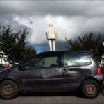 Statue on Old Car Art Installation in City Square Statue on old black car art installation in historic city square with dramatic sky.