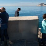 Tranquil Harbor View With Three People by the Sea Elderly couple and woman enjoy coastal harbor view with mountains, blue sky, and calm sea.