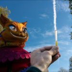 Whimsical Cheshire Cat Sculpture With Test Tube Experiment Hand holding test tube with golden liquid beneath colorful Cheshire cat sculpture outdoors.