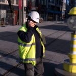 Construction Worker on Phone at Urban Job Site Construction worker on phone with excavator, safety vest, and urban street construction scene.