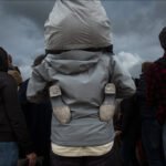 Man in Gray Jacket With Metallic Shapes Amid Crowd Crowd gathering with man in gray jacket featuring metallic shoe-like shapes on back.