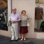 Stylish Elderly Couple Posing Outside Fashion Store Elderly couple posing stylishly outside clothing store with fashion sale sign.