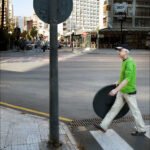 Elderly Man Carrying Table Across City Crosswalk Elderly man carrying table crossing city street with palm trees and modern buildings.