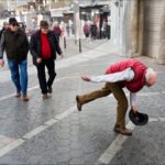 Elderly Man Playfully Picking Up Hat on Busy Street Elderly man picking up hat on lively European cobblestone street with pedestrians.