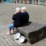 Elderly Women Sharing a Moment in Sunny Plaza Elderly women sitting on stone bench in sunny cobblestone plaza with murals and community vibe.