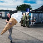 Man Enjoys Giant Ice Cream Cone at Outdoor Festival Man holding giant ice cream cone at outdoor festival with people and colorful tents.