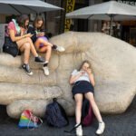 Girls on Giant Hand Sculpture in Urban Square Girls sitting on giant hand sculpture bench in busy urban square.