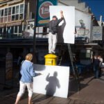 Man Balances on Ladder Installing Board in Busy Street Man balancing on ladder installing board on busy city street with cafes and people.
