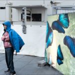Man in Blue Jacket Beside Vibrant Butterfly Banner Man in blue jacket standing by butterfly banner in urban industrial setting.