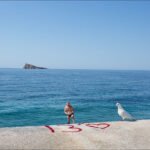 Man and Seagull by Peaceful Ocean Pier View Man standing on coastal pier with seagull, blue ocean waves, and rocky island view.