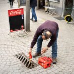 Man Places Coca-Cola Bottle by Cobblestone Street Drain Man placing Coca-Cola bottle near street drain on cobblestone urban sidewalk.