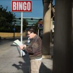 Man Reading Bingo Cards Under City Street Sign Man reading bingo cards on busy city street under bright bingo sign.
