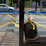 Elderly Man Waiting at City Bus Stop Elderly man sitting at urban bus stop bench with city street traffic and pedestrians.