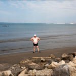 Man Relaxing on Sunny Beach with Ocean View Man standing on sandy beach with ocean waves and cloudy blue sky.
