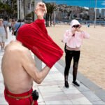 Elderly Man Playfully Struggles With Red Shirt at Beach Promenade Elderly man struggles with red shirt on busy beach promenade with people and palm trees.