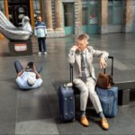 Traveler Waiting by Sculpture in Busy Airport Terminal Man waiting with luggage in airport terminal near modern sculpture and travelers.