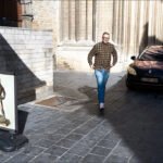 Man Walking on Sunlit Cobblestone Street with Car Man walking on historic cobblestone street with parked car and sunlight reflections.