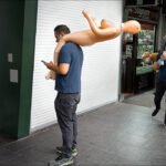Man Carrying Inflatable Doll on City Street Man carrying inflatable doll on busy city street near McDonald’s storefront.