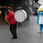 Musician and Cartoon Character Entertain Crowd in Train Station Musician performing with saxophone and drum beside cartoon character in busy train station.