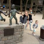 Children Playing by Bronze Statues in Playground Children playing near bronze statues in a playground with sand and climbing structures.