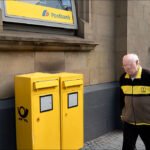 Elderly Man Passing Yellow Postbank Mailboxes Elderly man walking past yellow Postbank mailboxes outside historic bank building.