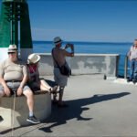 Sunny Seaside Promenade With Friends by Lighthouse Group enjoying sunny seaside promenade with ocean view and lighthouse backdrop.