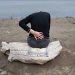 Contemplative Moment by Driftwood on Tranquil Shore Person sitting on driftwood by calm water, peaceful beach scene with introspective mood.
