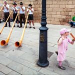 Young Girl Joins Alphorn Street Musicians in Lively Performance Young girl playing with alphorn street musicians in traditional alpine performance.