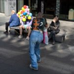 Colorful City Street Scene With Balloons and Shoppers Colorful city street scene with woman shopping, balloons, and people sitting on benches.