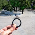 Framing City Life Through a Ring Perspective Hand holding ring framing urban square with cyclist, trees, kiosk, and parked bicycles.