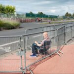 Man Sitting Behind Barricades on City Street Man sitting on chair behind street barricades in urban city with greenery and pedestrians.