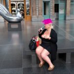 Woman in Black Dress Beside Modern Bird Sculpture Woman in black dress with pink fascinator sitting by modern bird sculpture indoors.