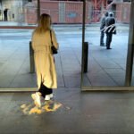 Woman in Trench Coat by Orange Spill in City Entrance Woman stepping in a yellow spill.