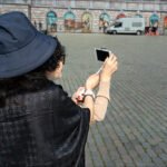 Woman Taking Selfie in Stylish Hat at Mural Square Woman taking selfie in cobblestone square with murals and stylish hat.