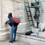 Young Musician with Guitar Case by Urban Statues Young musician with guitar case near urban bronze statues and stone wall.