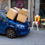 Blue car with stacked cardboard boxes and colorful mannequin on cobblestone urban street.