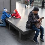 Children arm wrestling on stone bench in modern urban mall with adult reading nearby.