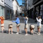 Young women posing playfully on a sunny cobblestone city street with modern buildings.