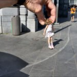 Playful optical illusion of a giant hand holding a tiny girl in a sunny city plaza.